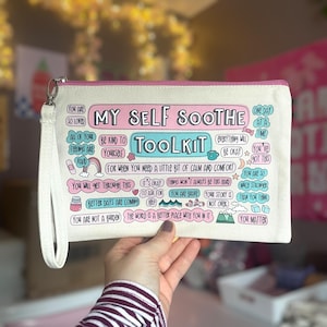 May include: A white canvas zippered pouch with a pink and blue design. The pouch has the words "My Self Soothe Toolkit" printed on it. The pouch is filled with positive affirmations and encouraging messages.