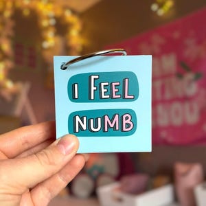 May include: A square light blue card held by a silver metal ring. The card features two teal rounded rectangular shapes containing the text I FEEL in white and pink letters on the top, and NUMB in white and pink letters on the bottom.