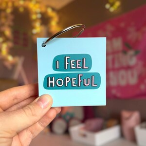 May include: A square light blue card with a metal binder ring in the top left corner. The card features two teal rounded rectangular shapes with the text "I FEEL HOPEFUL" printed in a white and pink sans-serif font with a black outline.