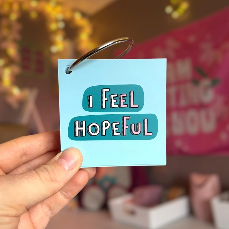 May include: A square light blue card with a metal binder ring in the top left corner. The card features two teal rounded rectangular shapes with the text "I FEEL HOPEFUL" printed in a white and pink sans-serif font with a black outline.