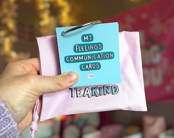 May include: A pink pouch with the text "TEAKIND" in white letters. The pouch contains a set of blue and white cards with the text "MY FEELINGS COMMUNICATION CARDS" and a silver ring.