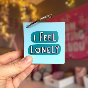 May include: A square light blue card held by a silver metal ring. The card features the text I FEEL LONELY in white and pink letters, set against two dark teal rounded rectangular shapes. The design is simple, clean, and centered.