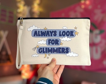 May include: Cream-colored canvas clutch with a black zipper and wrist strap. The clutch features the text "ALWAYS LOOK FOR GLIMMERS" in blue, surrounded by clouds, stars, and a crescent moon.