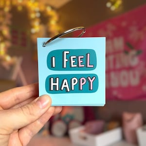 May include: A light blue square card held by a silver metal ring. The card features two dark teal horizontal rounded rectangles containing the white and pink text I FEEL HAPPY. The card is made of paper and has a hole punched in the top corner.