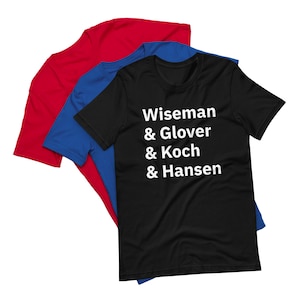 May include: Three short-sleeved t-shirts in red, blue, and black are layered. The black shirt features white text: "Wiseman & Glover & Koch & Hansen." The shirts are made of a soft material and are likely for casual wear.