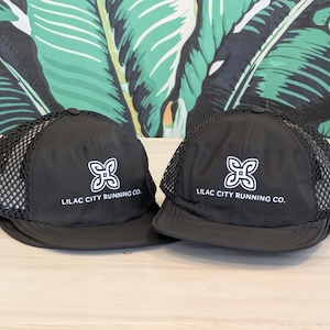 May include: Two black baseball caps with mesh netting on top. Each cap has a white logo and the text "LILAC CITY RUNNING CO." printed on the front. The caps are displayed on a light-colored wooden surface against a backdrop of green and white tropical leaves.
