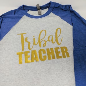 May include: A grey and blue baseball tee shirt with the words "Tribal Teacher" in gold glitter lettering.