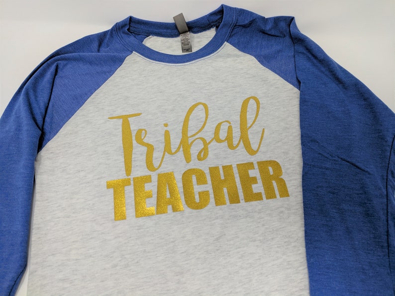 May include: A grey and blue baseball tee shirt with the words "Tribal Teacher" in gold glitter lettering.