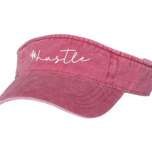 May include: A red, distressed, cotton visor with a white embroidered hashtag and the word "hustle".