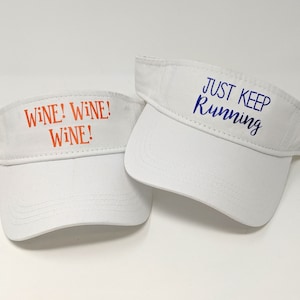 May include: Two white visor hats with different text. One hat says "Wine! Wine! Wine!" in orange text. The other hat says "Just Keep Running" in blue text.
