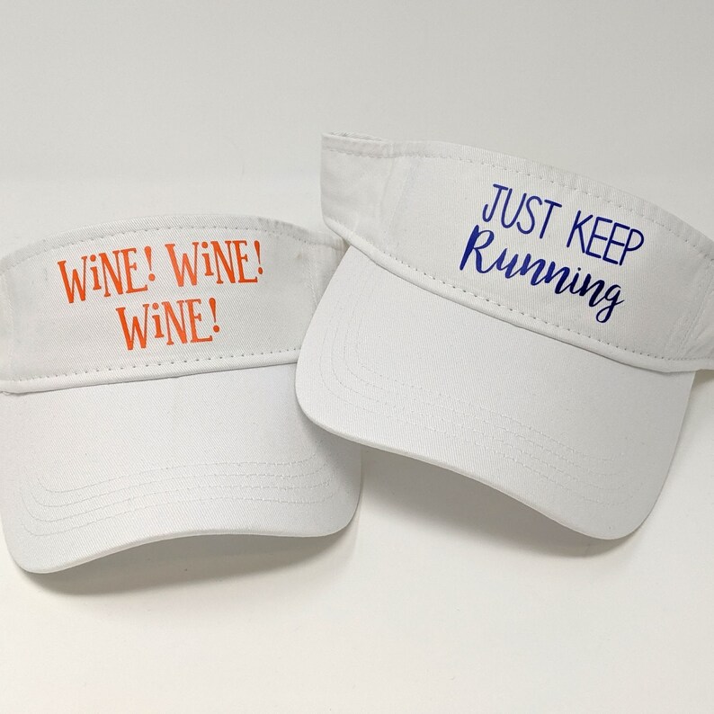 May include: Two white visor hats with different text. One hat says "Wine! Wine! Wine!" in orange text. The other hat says "Just Keep Running" in blue text.