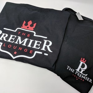 Pu&ograve; includere: Due magliette nere con un logo bianco che dice "The Premier Lounge" e una corona rossa. Una maglietta ha un logo pi&ugrave; grande con una stella e l'altra ha un logo pi&ugrave; piccolo con un "PL" stilizzato al centro.