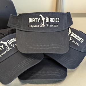 May include: A stack of four navy blue visors with white embroidered text that reads "Dirty Birdies" and "Hollywood Open" with a golf club and ball graphic. The text also reads "Est. 2023".