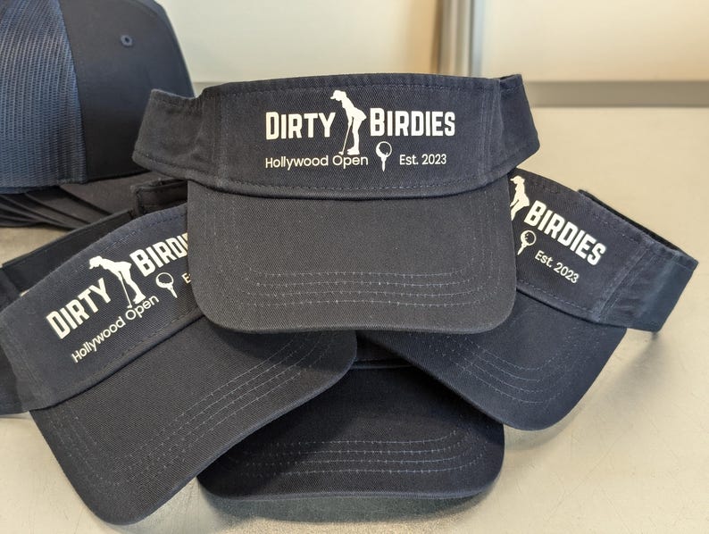 May include: A stack of four navy blue visors with white embroidered text that reads "Dirty Birdies" and "Hollywood Open" with a golf club and ball graphic. The text also reads "Est. 2023".