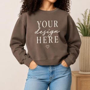 May include: A dark brown crewneck sweatshirt featuring the white text "YOUR design HERE" centered on the chest, positioned above a small white heart outline. The sweatshirt has long sleeves with ribbed cuffs and a ribbed hem, paired with blue denim jeans.