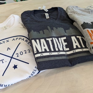 K&ouml;nnte beinhalten: Drei gefaltete T-Shirts mit dem Text "NATIVE ATX APPAREL" und einer Skyline-Grafik. Das wei&szlig;e Shirt hat ein marineblaues Logo mit dem Text "EST. 2022" und einen Stern. Das graue Shirt hat einen orangen Streifen und den Text "NATIVE APPAREL". Das dunkelblaue Shirt hat eine graue Skyline-Grafik und den Text "NATIVE ATX APPAREL".