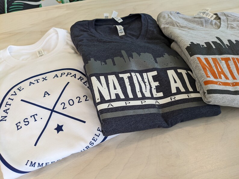 K&ouml;nnte beinhalten: Drei gefaltete T-Shirts mit dem Text "NATIVE ATX APPAREL" und einer Skyline-Grafik. Das wei&szlig;e Shirt hat ein marineblaues Logo mit dem Text "EST. 2022" und einen Stern. Das graue Shirt hat einen orangen Streifen und den Text "NATIVE APPAREL". Das dunkelblaue Shirt hat eine graue Skyline-Grafik und den Text "NATIVE ATX APPAREL".