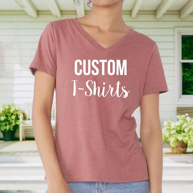 May include: A woman is wearing a dusty rose coloured v-neck t-shirt with the text "CUSTOM T-Shirts" printed in white on the front.