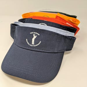 May include: A navy blue golf visor with a white embroidered logo that says "Troy Durham Memorial Golf Tournament" and a silhouette of a golfer swinging a club.