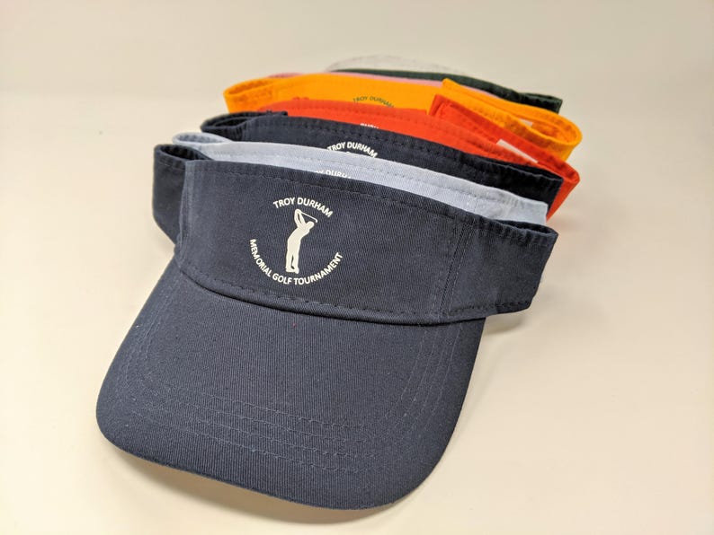 May include: A navy blue golf visor with a white embroidered logo that says "Troy Durham Memorial Golf Tournament" and a silhouette of a golfer swinging a club.