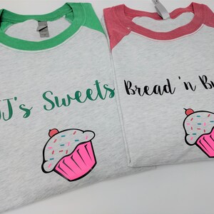May include: Two grey baseball t-shirts with green and red sleeves. One shirt says "JJ's Sweets" and the other says "Bread 'n Butter". Both shirts have a pink cupcake with sprinkles on them.