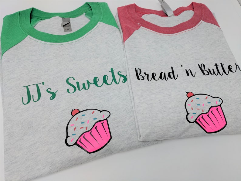 May include: Two grey baseball t-shirts with green and red sleeves. One shirt says "JJ's Sweets" and the other says "Bread 'n Butter". Both shirts have a pink cupcake with sprinkles on them.