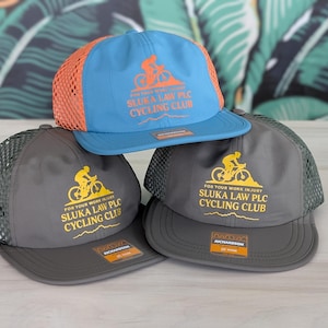 May include: Three baseball caps with mesh backs are stacked. The top cap is blue and orange, while the other two are gray. Each cap features the text "SLUKA LAW PLC CYCLING CLUB" and a cyclist graphic in yellow.