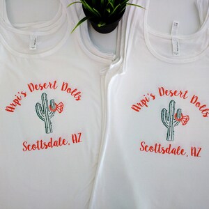 May include: Two white tank tops with a green cactus graphic and the text "Arpi's Desert Dolls Scottsdale, AZ" in red.