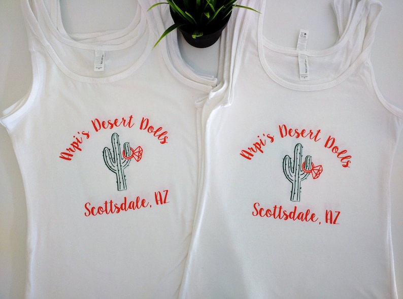 May include: Two white tank tops with a green cactus graphic and the text "Arpi's Desert Dolls Scottsdale, AZ" in red.