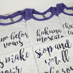 May include: Four gray t-shirts with purple sleeves and black text. The text on the shirts reads "wine before vows", "hakuna moscat", "stop and smell the ros&eacute;", and "champs,  camp,  and  ros&eacute;".