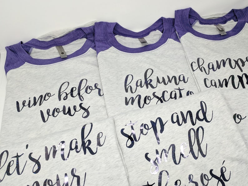 May include: Four gray t-shirts with purple sleeves and black text. The text on the shirts reads "wine before vows", "hakuna moscat", "stop and smell the ros&eacute;", and "champs,  camp,  and  ros&eacute;".