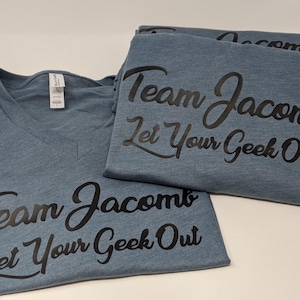 May include: Three blue t-shirts with a black graphic that says "Team Jacomb Let Your Geek Out".