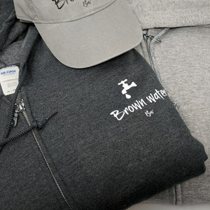 May include: Two hooded sweatshirts with a zipper closure. The sweatshirts are gray and dark gray. Both sweatshirts have a white logo with a faucet and the text "Brown Water Brew". A gray baseball cap with the same logo is also pictured.