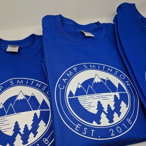K&ouml;nnte beinhalten: Drei k&ouml;nigsblaue T-Shirts mit einem wei&szlig;en kreisf&ouml;rmigen Logo, das eine Bergkette, einen See und Kiefern zeigt. Das Logo lautet "Camp Smithson Est. 2018".
