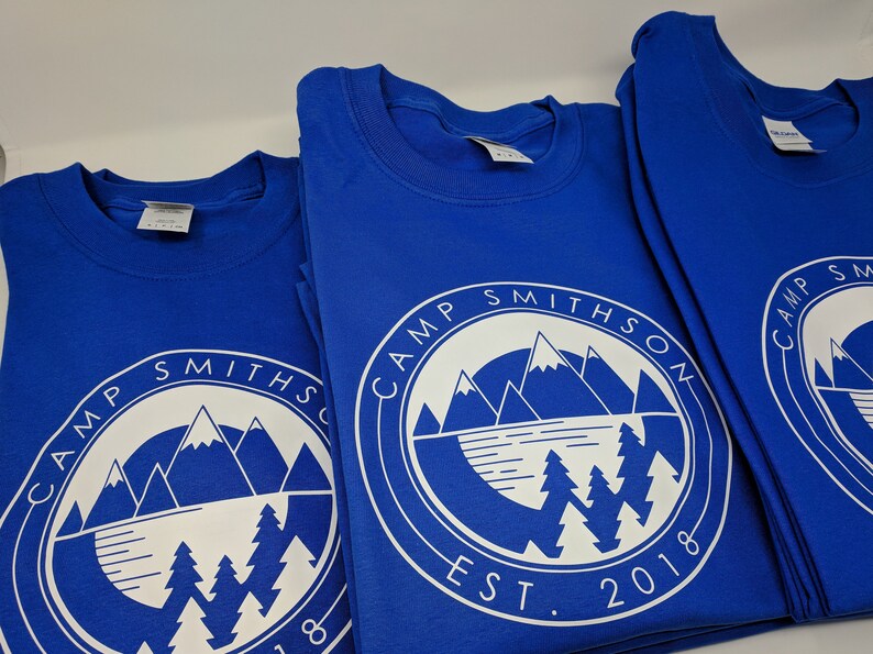 K&ouml;nnte beinhalten: Drei k&ouml;nigsblaue T-Shirts mit einem wei&szlig;en kreisf&ouml;rmigen Logo, das eine Bergkette, einen See und Kiefern zeigt. Das Logo lautet "Camp Smithson Est. 2018".