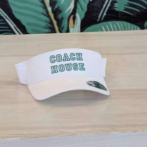 Może przedstawiać: Biały daszek z napisem "COACH HOUSE" w zielonych blokowych literach. Daszek ma zakrzywiony daszek i biały regulowany pasek. Daszek jest wykonany z lekkiego materiału, odpowiedniego do aktywności na świeżym powietrzu.