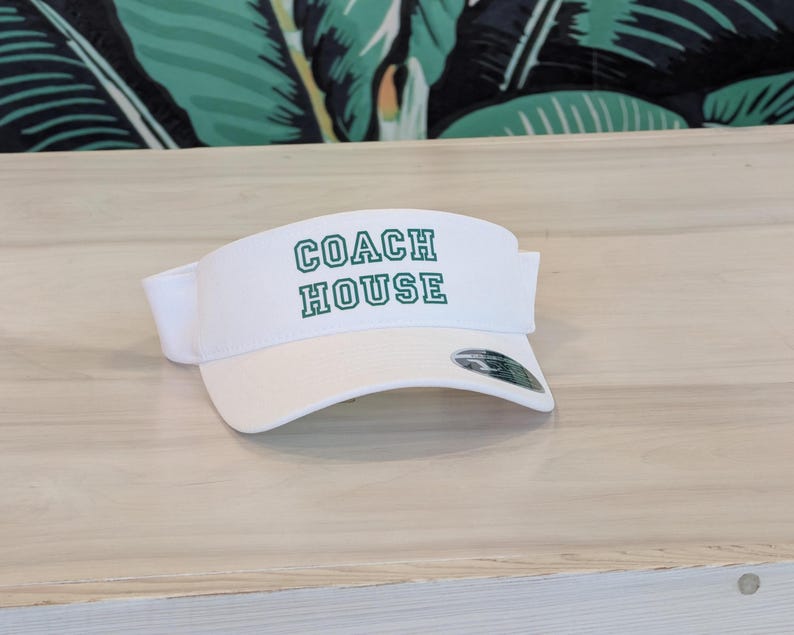 Może przedstawiać: Biały daszek z napisem "COACH HOUSE" w zielonych blokowych literach. Daszek ma zakrzywiony daszek i biały regulowany pasek. Daszek jest wykonany z lekkiego materiału, odpowiedniego do aktywności na świeżym powietrzu.
