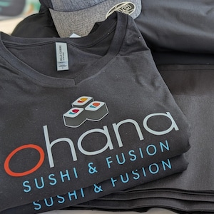May include: A stack of black t-shirts with the text "ohana sushi & fusion" printed on them. The shirts are folded and stacked on a wooden surface.