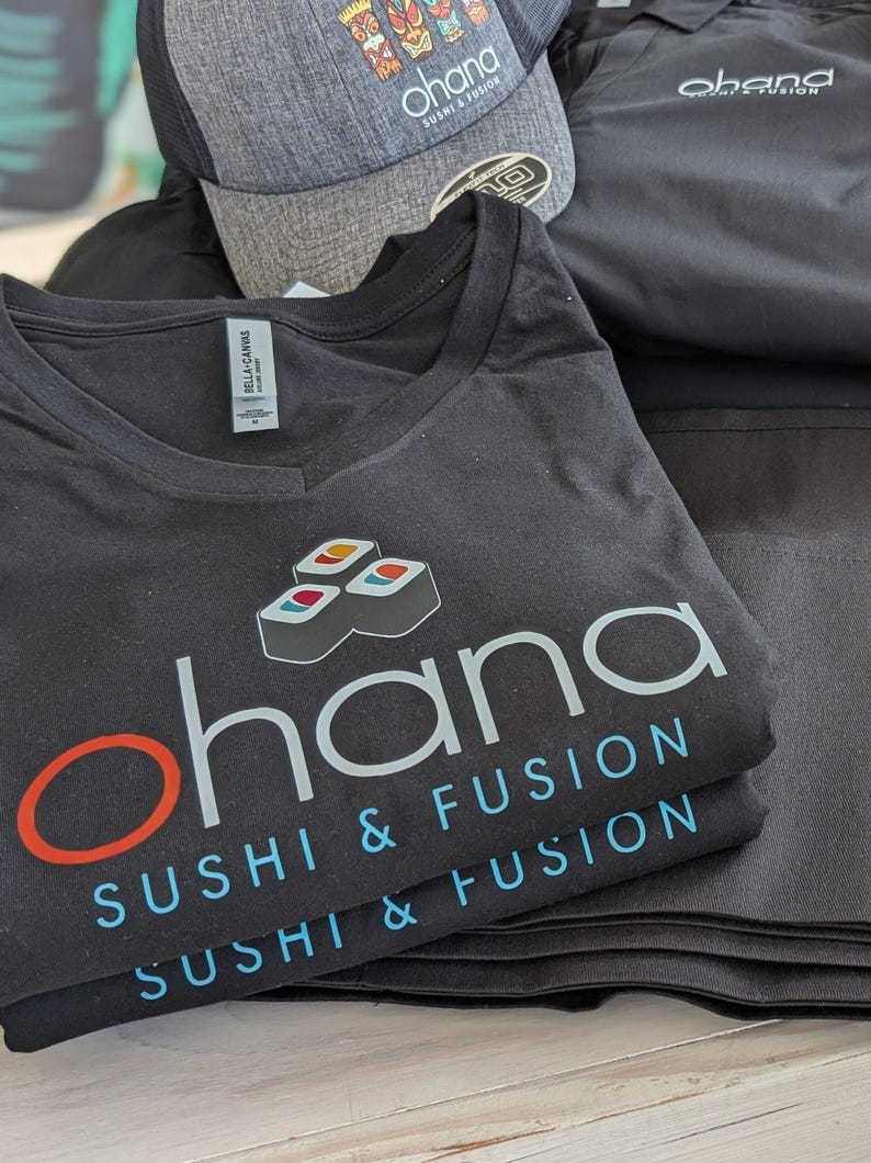 May include: A stack of black t-shirts with the text "ohana sushi & fusion" printed on them. The shirts are folded and stacked on a wooden surface.