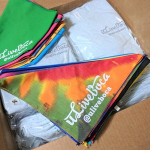 May include: A cardboard box filled with a variety of merchandise, including tie-dye bandanas, plain t-shirts, and solid color bandanas. The bandanas feature the text "uLiveBoca @uliveboca".