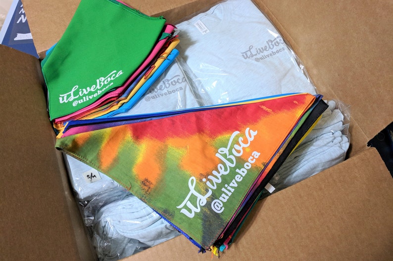 May include: A cardboard box filled with a variety of merchandise, including tie-dye bandanas, plain t-shirts, and solid color bandanas. The bandanas feature the text "uLiveBoca @uliveboca".