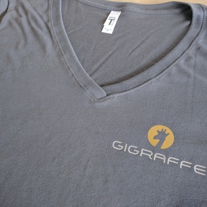 Pu&ograve; includere: Maglietta grigia a collo a V con un logo circolare giallo che presenta una silhouette di giraffa e il testo "GIGRAFFE" in bianco.