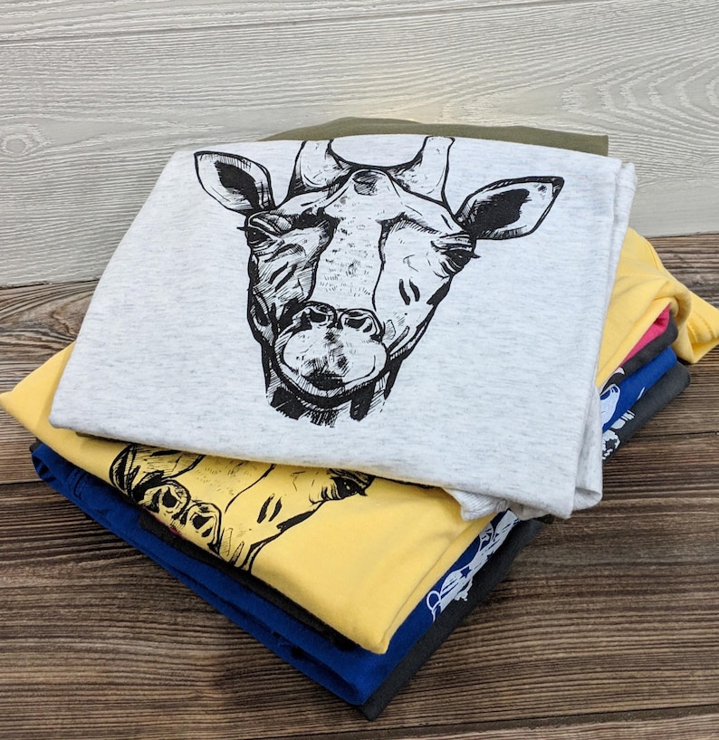 Pu&ograve; includere: Una pila di magliette con una grafica di giraffa stampata sulla maglietta superiore. Le magliette sono di vari colori, tra cui grigio, giallo, blu, rosa e verde.