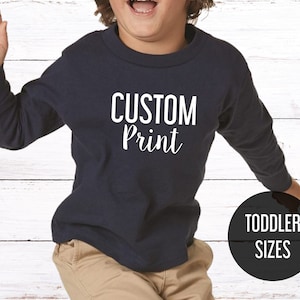 May include: A navy blue long-sleeved toddler shirt with the text "CUSTOM Print" in white. The shirt is shown on a child model. The text "TODDLER SIZES" is in a black circle in the lower right corner of the image.