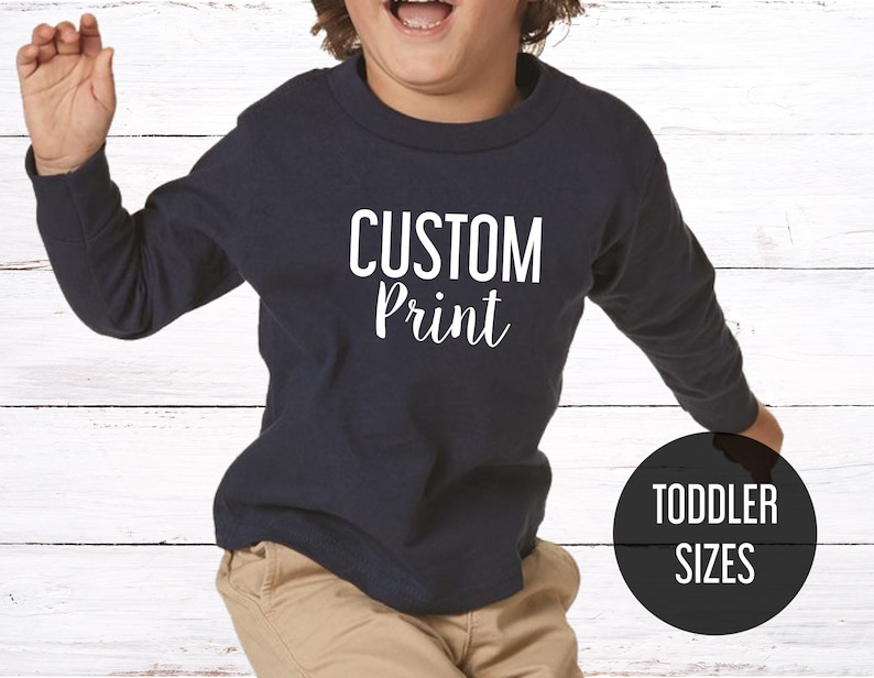 May include: A navy blue long-sleeved toddler shirt with the text "CUSTOM Print" in white. The shirt is shown on a child model. The text "TODDLER SIZES" is in a black circle in the lower right corner of the image.