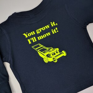 May include: A navy blue long-sleeved shirt with the text "You grow it, I'll mow it!" in yellow lettering. A yellow silhouette of a lawnmower is below the text.