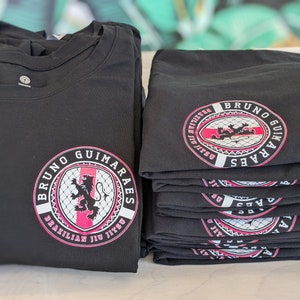 K&ouml;nnte beinhalten: Stapel schwarzer T-Shirts mit einem pinkfarbenen und wei&szlig;en Logo, auf dem "Bruno Guimaraes Brazilian Jiu Jitsu" steht.