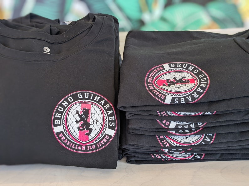 K&ouml;nnte beinhalten: Stapel schwarzer T-Shirts mit einem pinkfarbenen und wei&szlig;en Logo, auf dem "Bruno Guimaraes Brazilian Jiu Jitsu" steht.