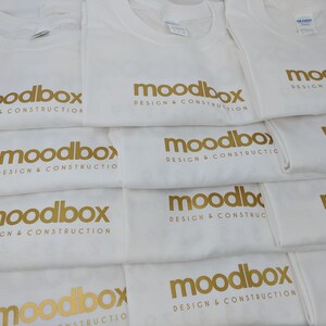 K&ouml;nnte beinhalten: Ein Stapel wei&szlig;er T-Shirts mit dem goldenen Text "moodbox design & construction" darauf gedruckt.