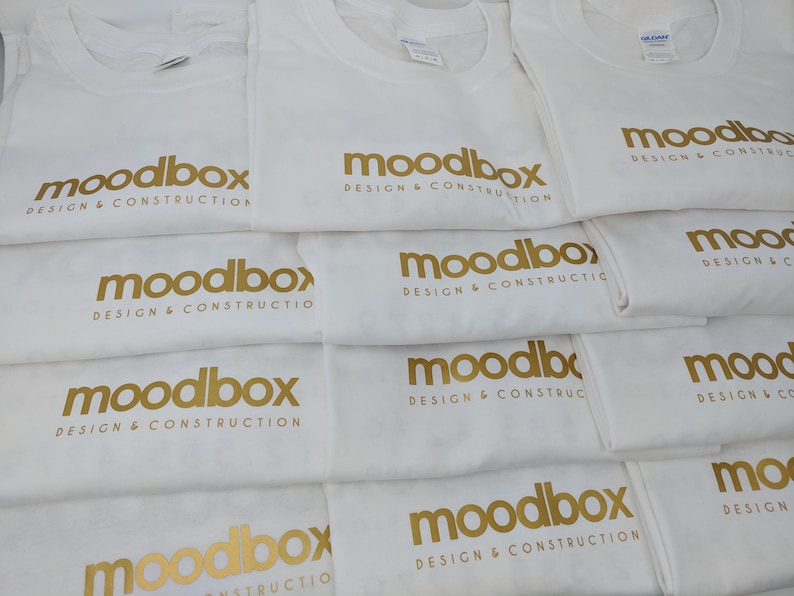K&ouml;nnte beinhalten: Ein Stapel wei&szlig;er T-Shirts mit dem goldenen Text "moodbox design & construction" darauf gedruckt.